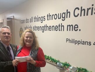 Sheriff Jody Greene presents a donation to community CPR's Christmas relief efforts. the Bible verse in question is on the wall in the background, and has been used in numerous photographs.
