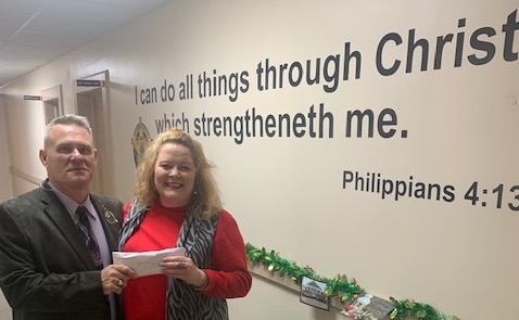 Sheriff Jody Greene presents a donation to community CPR's Christmas relief efforts. the Bible verse in question is on the wall in the background, and has been used in numerous photographs.